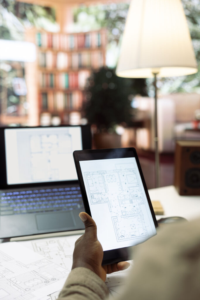 Hand mit Tablet zeigt technischen Gebäudegrundriss in moderner Planungsumgebung mit Laptop und Bauunterlagen