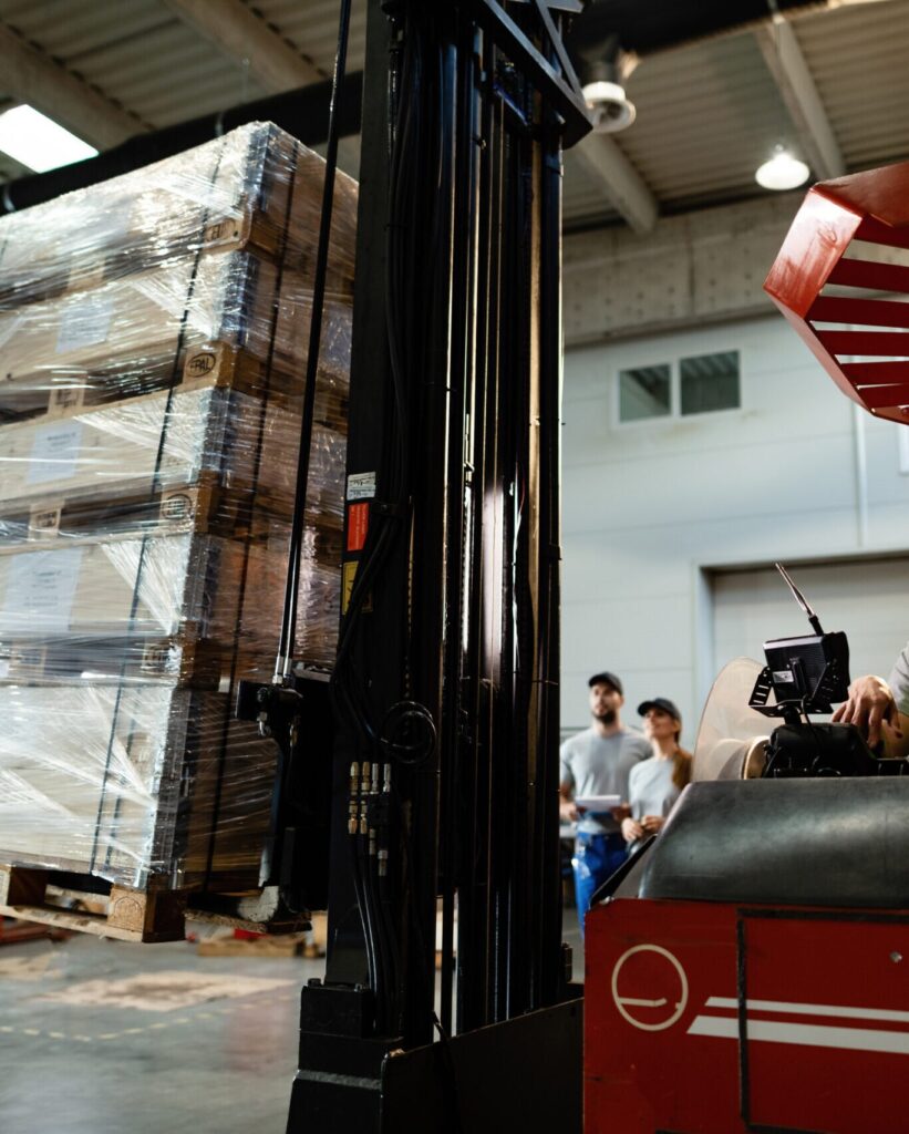 Gabelstapler hebt palettierte Ware in einer Industriehalle, im Hintergrund stehen zwei Mitarbeitende