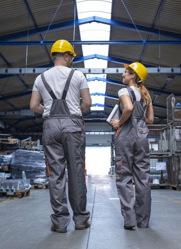Zwei Personen in grauen Overalls und gelben Helmen gehen nebeneinander durch eine große Industriehalle